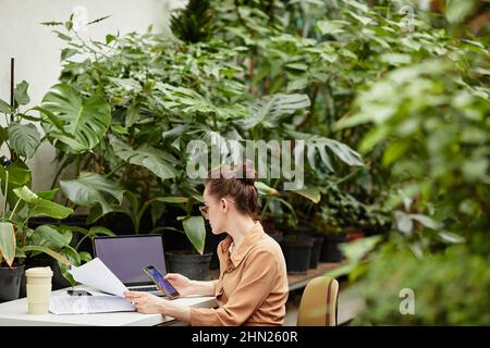 Young businesswoman scrolling in smartphone while checking papers and answering questions of clients online by workplace Stock Photo