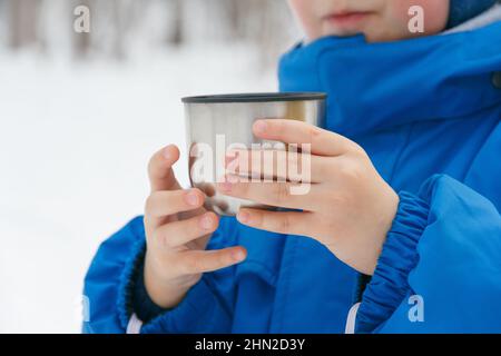 Child hands holds a cup. Child drinking a cup of tea on the street while walking. A cup of hot drink from a thermos outside in winter. Winter walk Stock Photo