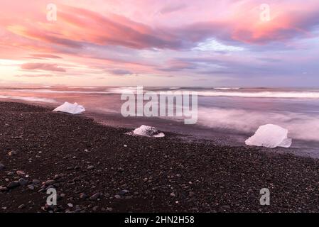 Blocks of ice on a black beach under dramatic sky glowing pink and orange in midnight sun light in summer