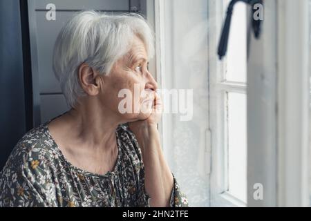 An elderly woman pensioner looks out the window sitting in his house. High quality photo Stock Photo