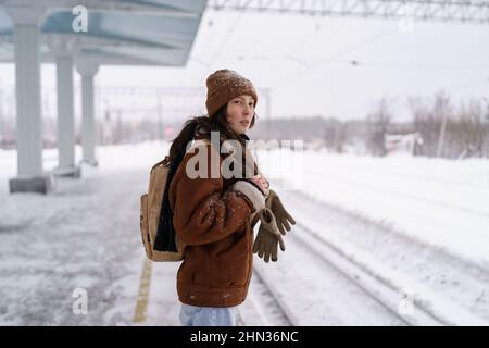 Young pensive Asian woman with backpack standing at snow-covered railway platform in winter Stock Photo