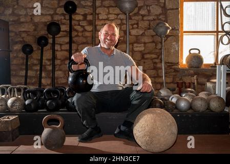 Luxemburg, Luxembourg. 09th Feb, 2021. Georges Christen squats in front of historic dumbbells and weights in his training barn. The Luxembourger is one of the strongest men in the world: he has 26 entries in the Guinness Book of Records. In just a few seconds, he tears up telephone books or bends thick nails. (to dpa: Georges Christen: The man who rolls up pans) Credit: Harald Tittel/dpa/Alamy Live News Stock Photo