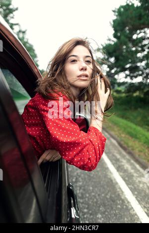 A girl in a light red airy dress, a leather jacket in a rockstyle on an empty road in a summer forest Stock Photo