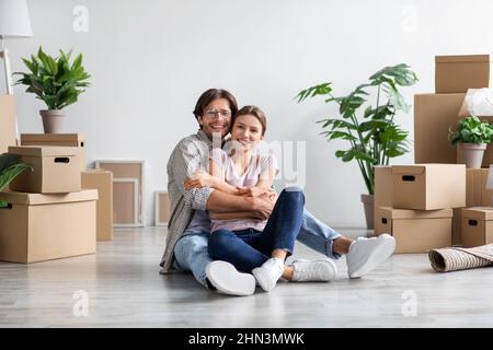 Satisfied european young guy in glasses in casual hugging female, sit on floor with cardboard boxes Stock Photo