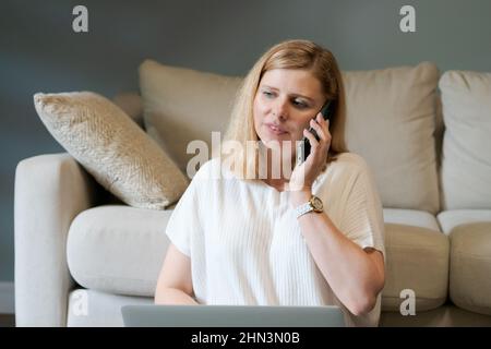 Pretty woman working remotely from home, freelance business woman sitting on floor near sofa with laptop, talking on phone, taking notes in laptop in casual clothes in living room Stock Photo