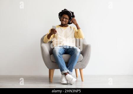 Modern Technologies. Black Millennial Guy With Smartphone And Headphones Relaxing In Chair Stock Photo