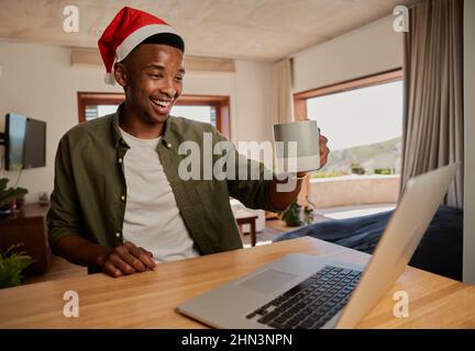 Young adult black male wearing Christmas hat, drinking coffee while on online call. Sitting at kitchen counter using laptop. Stock Photo