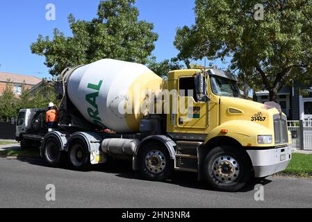 Yellow Boral cement mixer truck parked on a sunny suburban street, with a man in fluorescent work wear sitting at the truck's rear Stock Photo
