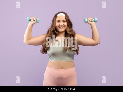 Happy pretty young european plus size female in uniform doing hand exercises with dumbbells