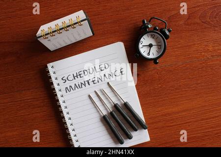 Top view image of calendar, alarm clock, screwdriver and book with text SCHEDULED MAINTENANCE. Stock Photo