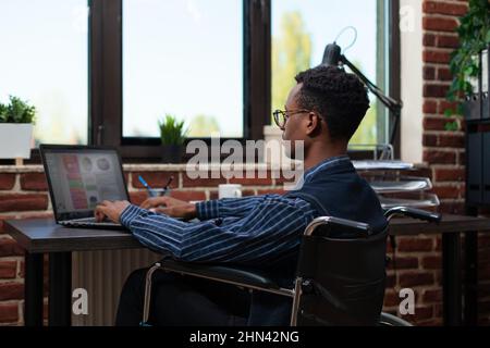 Startup employee who uses wheelchair working on laptop with business analytics in red brick office. African american marketing specialist analyzing key performance indicators. Stock Photo