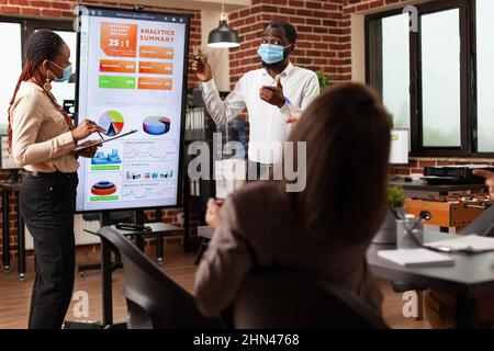 Businesspeople with medical face mask to prevent infection with coronavirus showing business graphs discussing company strategy in startup office. Diverse people working at financial presentation Stock Photo