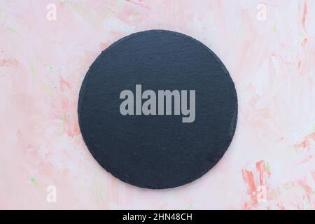Round black slate plate on pink spring background. Kitchen stone tray for serving food. Black granite circle board. Empty space for menu or recipe. Top view copy space, mockup Stock Photo