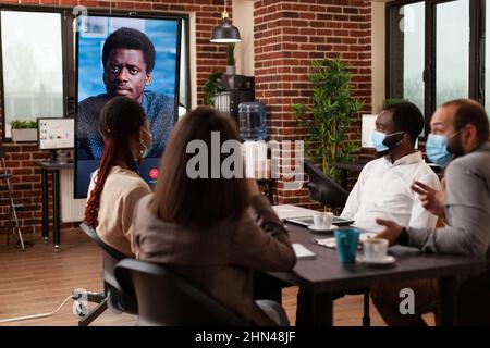 Multi-ethnic group of entrepreneurs with protective face mask against covid19 discussing management strategy with remote collegue. Online videocall meeting conference in startup company office Stock Photo