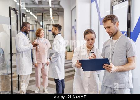 Two young confident healthcare workers discussing medical documentation against three communicating colleagues in lab coats Stock Photo