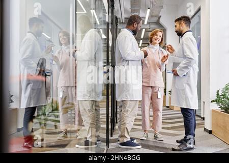 Two intercultural male healthcare workers and their female colleague having talk in corridor of contemporary medical center Stock Photo