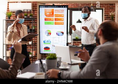 African american managers showing marketing presentation explaining business graphs working at company partnership in startup office. Businessteam with medical face mask against covid19 Stock Photo