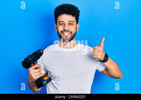 Young arab man with beard using drill smiling happy and positive, thumb up doing excellent and approval sign Stock Photo