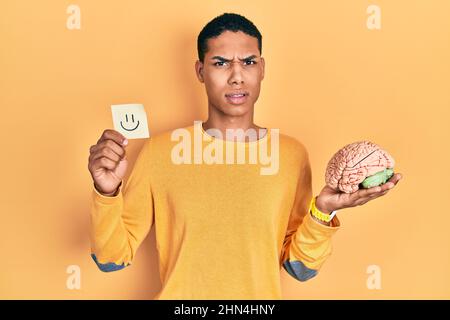 Young african american guy holding smile reminder and brain clueless and confused expression. doubt concept. Stock Photo