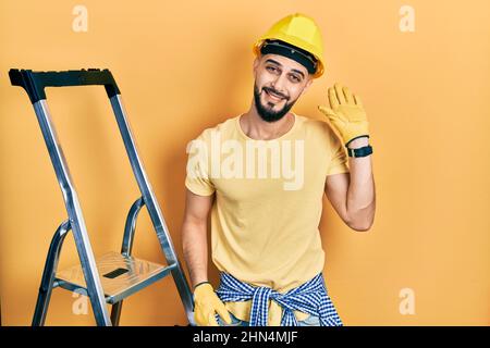 Handsome man with beard by construction stairs wearing hardhat waiving saying hello happy and smiling, friendly welcome gesture Stock Photo