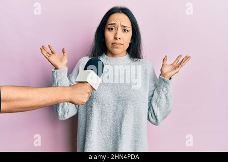 Young hispanic girl being interviewed by reporter holding microphone clueless and confused with open arms, no idea and doubtful face. Stock Photo