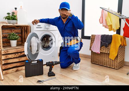 Young indian technician working on washing machine covering eyes with hand, looking serious and sad. sightless, hiding and rejection concept Stock Photo