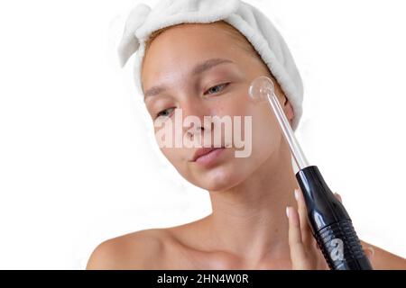 Isolated portrait of caucasian young woman without make up with acne and band on head holding darsonval on face looking down on white background Stock Photo