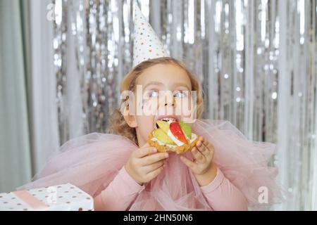 Pretty little girl with short fair curly hair and stars on face in pink poofy dress and birthday cap eating fruit cake. Stock Photo