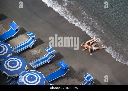 Two bathers sitting at the waters edge with blue sunbeds and umbrellas behind them Stock Photo