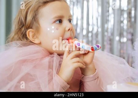 Portrait of pretty little girl with short fair curly hair and golden stars on face in pink poofy dress blow noisemaker. Stock Photo