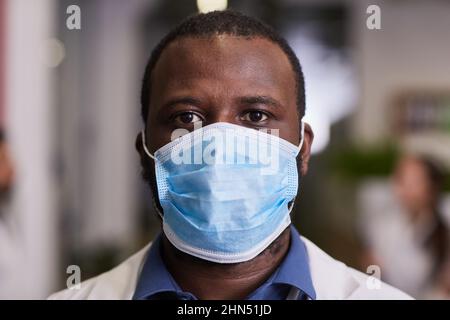 Young serious African-American healthcare worker in protective mask and workwear standing against hospital interior and looking at camera Stock Photo