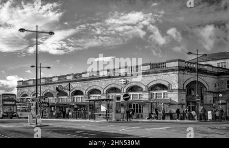 The external view of a railway station.  The 19th Century  building has bus stops at its front and people stand and sit waiting for buses. Stock Photo