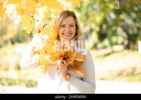 Attractive smiling middle-aged woman with long wavy fair hair in roll-neck sweater hold bouquet of fallen yellow leaves. Stock Photo