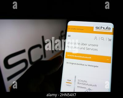 Person holding smartphone with weboage of German credit protection company Schufa Holding AG on screen with logo. Focus on center of phone display. Stock Photo