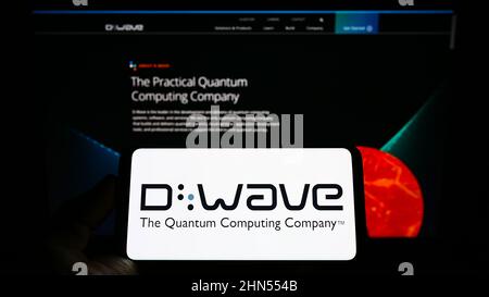 Person holding mobile phone with logo of Canadian hardware company D-Wave Systems Inc. on screen in front of web page. Focus on phone display. Stock Photo