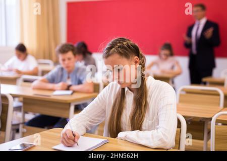 Teen girl listening to lecturer and writing in notebook in classroom Stock Photo