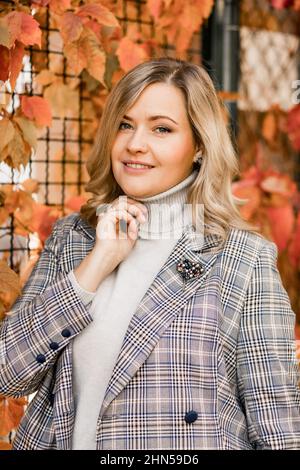 Smiling middle-aged woman with long hair in checkered jacket and grey roll-neck sweater posing touching neck with hand. Stock Photo