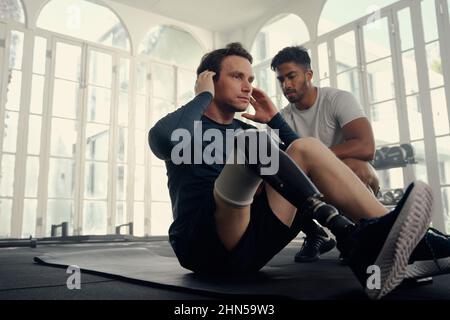 Paralympic athlete training with his coach in the gym . Man with prosthetic leg doing sit-ups in the gym with his coach Stock Photo