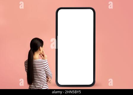 Back view of woman looking at big smartphone with blank white screen and thinking, device with space for mockup Stock Photo