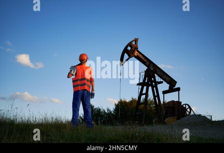 Rear view of oilman in orange uniform with working tool who watching at pump oil working in blue sky background. Petroleum extraction machine in energetic industry. Stock Photo