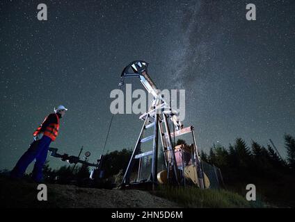 Bottom view of man in workwear standing to the left of oil pump and watching for its activities. Mechanic controls efficient night work of oil drilling rig. Starry sky and forest on background. Stock Photo