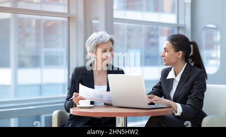 Two women analyzing documents while sitting on a table in office. Woman executives at work in office discussing some paperwork using laptop Stock Photo