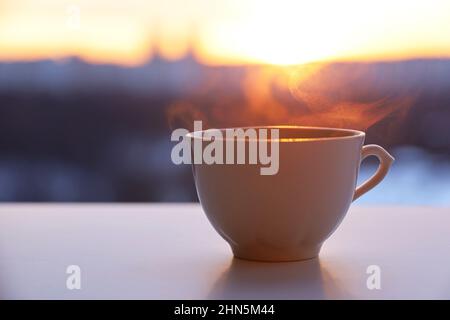 Hot coffee cup with smoke on sunshine background. View from the window to silhouettes of city buildings Stock Photo