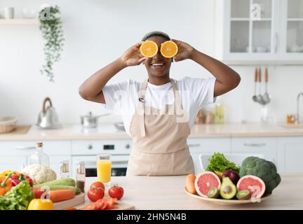 Glad funny millennial black female in apron have fun, puts orange slices to eyes at table with colorful vegetables Stock Photo