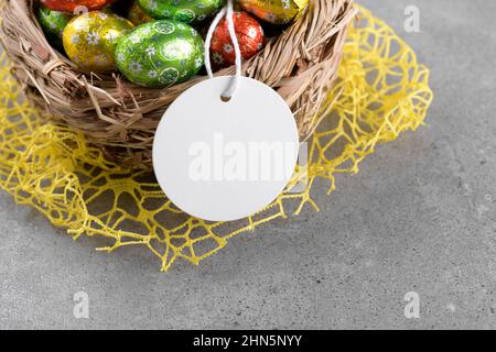Round white Easter tag mockup with Happy Easter chocolate easter eggs in birds nest on grey stone background. Easter gift tag or sign for message gree Stock Photo