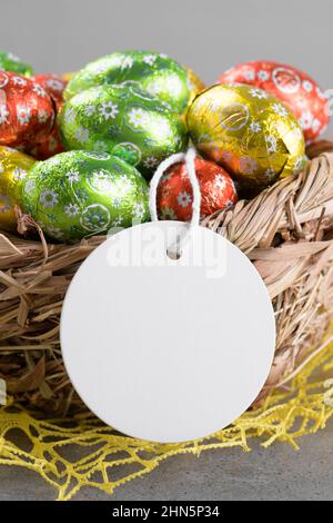 Round white Easter tag mockup with Happy Easter chocolate easter eggs in birds nest on grey stone background. Easter gift tag or sign for message gree Stock Photo