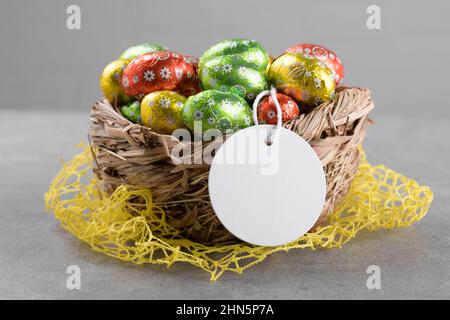 Round white Easter tag mockup with Happy Easter chocolate easter eggs in birds nest on grey stone background. Easter gift tag or sign for message gree Stock Photo