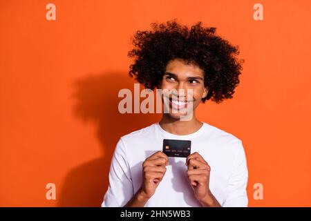 Photo of pretty charming guy wear white shirt holding bank card looking empty space isolated orange color background Stock Photo