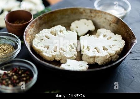 Cauliflower steak cooking. Raw cut cauliflower lie in a frying pan. Olive oil, herbs, various spices nearby. Dark background. Place for text. Stock Photo
