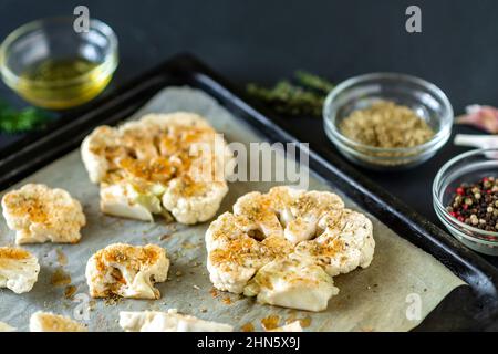 Cauliflower steak cooking. Raw cauliflower sprinkled with spices lies on a baking sheet. Olive oil, herbs, various spices nearby. Dark background. Stock Photo